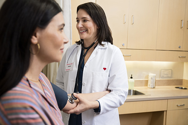 A healthcare professional wearing a white lab coat and stethoscope smiles while taking the blood pressure of a seated patient in a medical examination room. The room has wooden cabinets and countertops in the background.