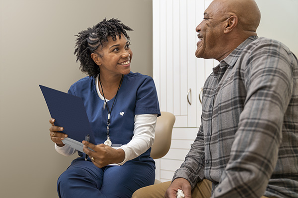 A female healthcare provider in blue scrubs is smiling and holding a clipboard while interacting with a male patient in a plaid shirt. They appear to be having a pleasant conversation in a medical office setting.