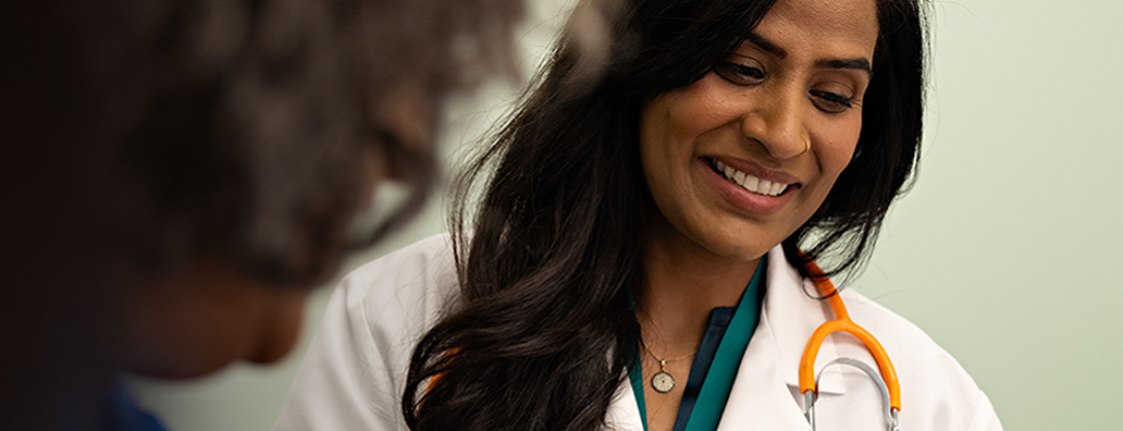 A female pharmacist in a white coat is smiling and giving a high-five to a young girl. The girl's mother, wearing a gray sweater, stands next to her, smiling. They are in a pharmacy with a healthcare-themed wallpaper in the background.