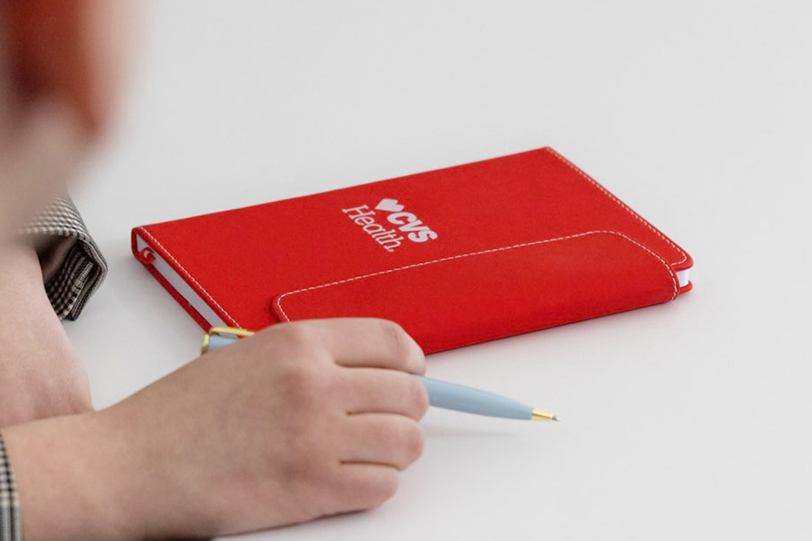 A person holds a light blue pen near a red notebook that reads "CVS Health" in white text. The notebook is placed on a white surface. The person’s face is not visible, but part of their hand and arm, dressed in a checked sleeve, is shown in the foreground.