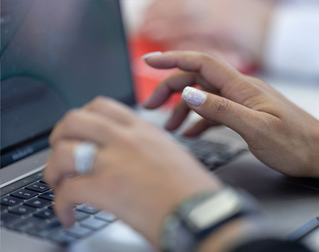 Close-up of a person's hands typing on a laptop keyboard. The person has manicured nails with sparkly nail polish, and is wearing a ring and a watch. Another person's blurred hand is visible in the background.