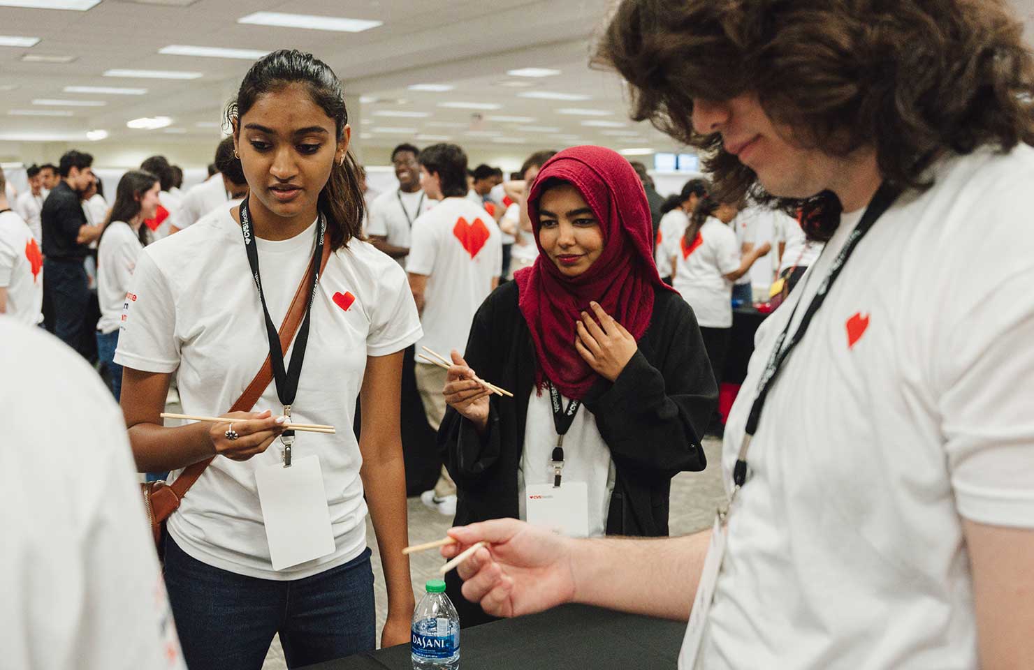 A diverse group of people stand together in a room wearing matching white t-shirts with red heart logos. They are actively engaged in a discussion, with some holding and examining objects. The background shows more people and a bright, well-lit space.