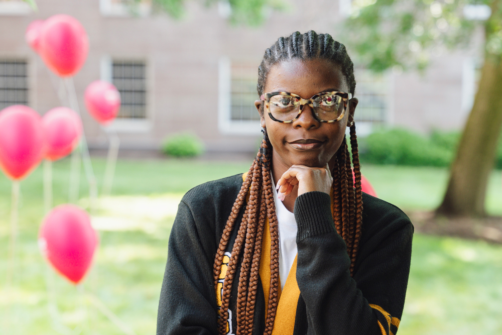 A person with long braids and glasses stands outdoors with a slight smile, resting their chin on their hand. They wear a black and yellow jacket, and red balloons are visible in the background, with a building and greenery behind them.