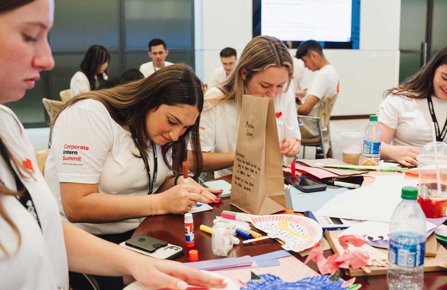 A group of interns in white shirts, labeled "Corporate Intern Summit," sit at a table engaging in an arts and crafts activity. They are surrounded by colorful paper, glue sticks, and other supplies. One intern focuses intently on their craft project.