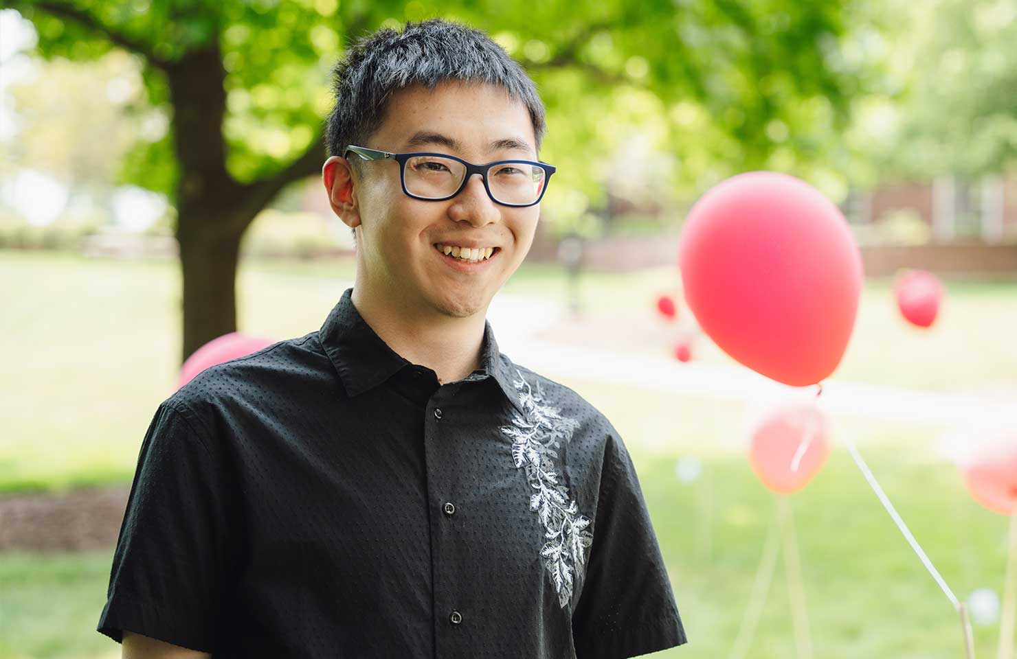 A person wearing glasses and a black shirt with white embroidery stands outdoors, smiling. Behind them, red balloons are tied to strings, with a tree and grassy area in the background.