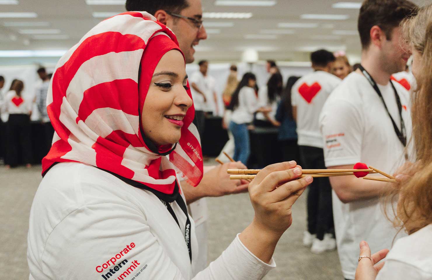 A woman wearing a red and white striped hijab and a white T-shirt with "Corporate Internship" written on the sleeve uses chopsticks to place a small pom-pom on another person's hand. The background shows several other people engaged in similar activities.