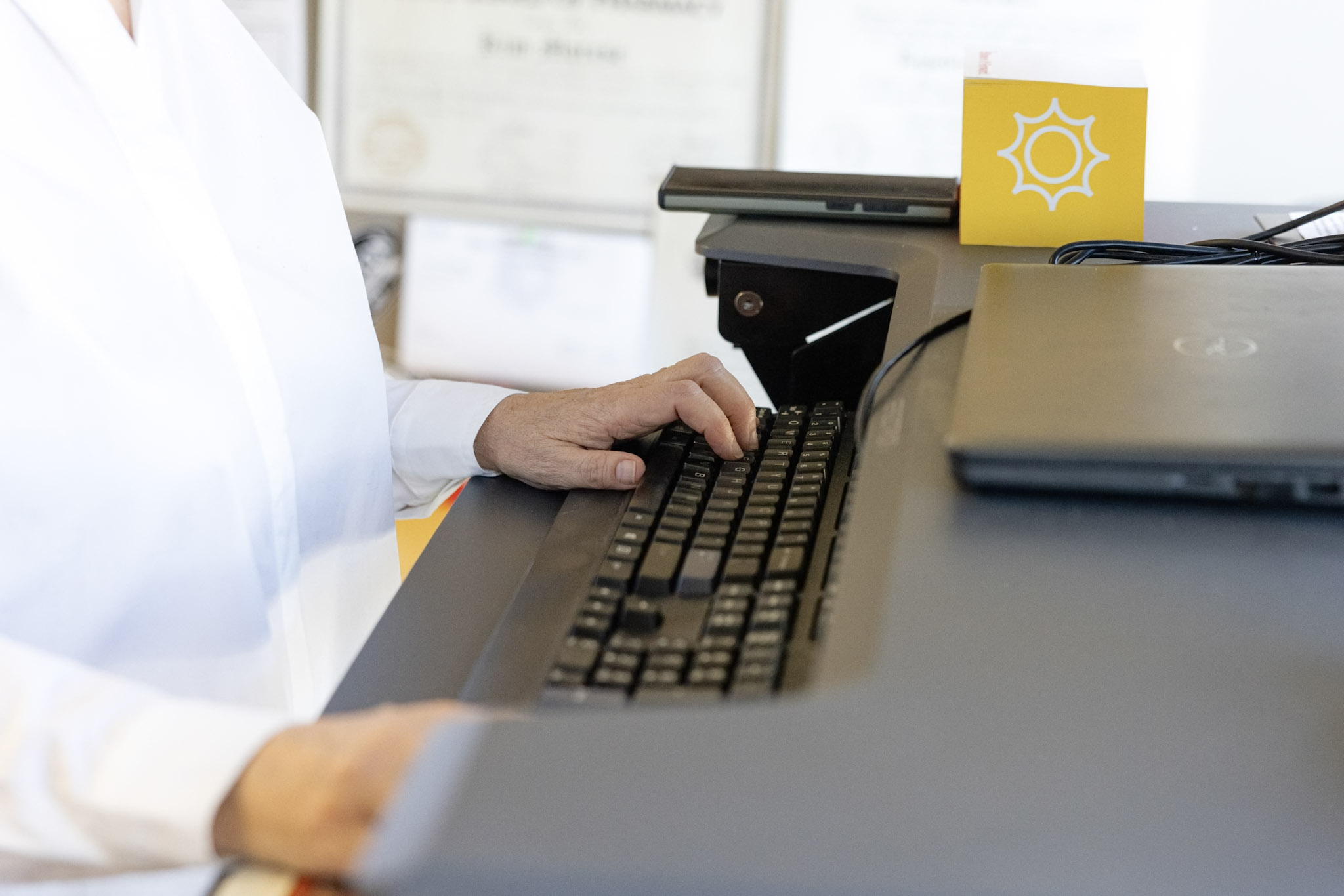 Person wearing a white shirt, typing on a keyboard at a standing desk with a laptop and a yellow card featuring a sun icon. Certificates are slightly visible in the blurry background.