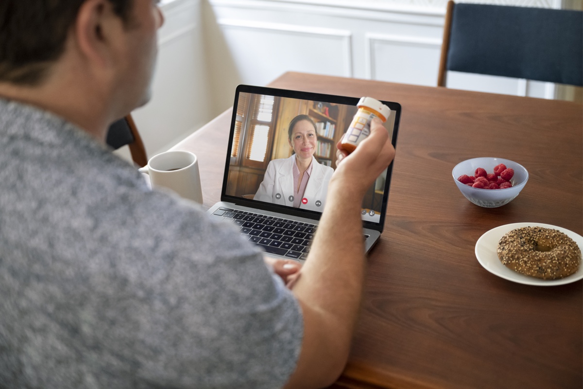 A person sitting at a table having a video call with a healthcare professional on a laptop. The person is holding a prescription bottle, and the table has a cup, a bowl of raspberries, and a plate with a bagel.