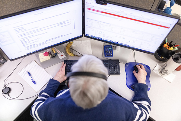 A person wearing headphones is seated at a desk with two computer monitors displaying information. The desk has various items including a notepad, pen, calculator, tissue, and a drink bottle. The individual is typing on the keyboard and using a mouse.