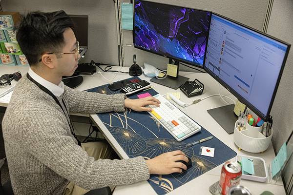 Person sitting at a desk using a computer with dual monitors. They are typing on the keyboard and holding a mouse. The desk has various items including sticky notes, a can of soda, and office supplies. The background features office cubicle decor.