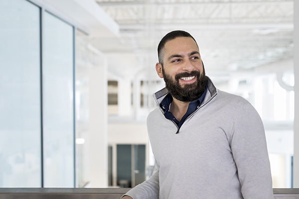 A smiling man with a beard and short hair stands indoors with a modern office in the background. He is wearing a light grey sweater over a dark collared shirt and is looking to the side. The atmosphere is bright and clean with large glass panels and white walls.