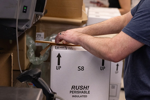 A person handling a shipping label and placing it onto a white box labeled "RUSH! PERISHABLE INSULATED" with arrows indicating the correct orientation. Various other boxes and packing materials are visible in the background.