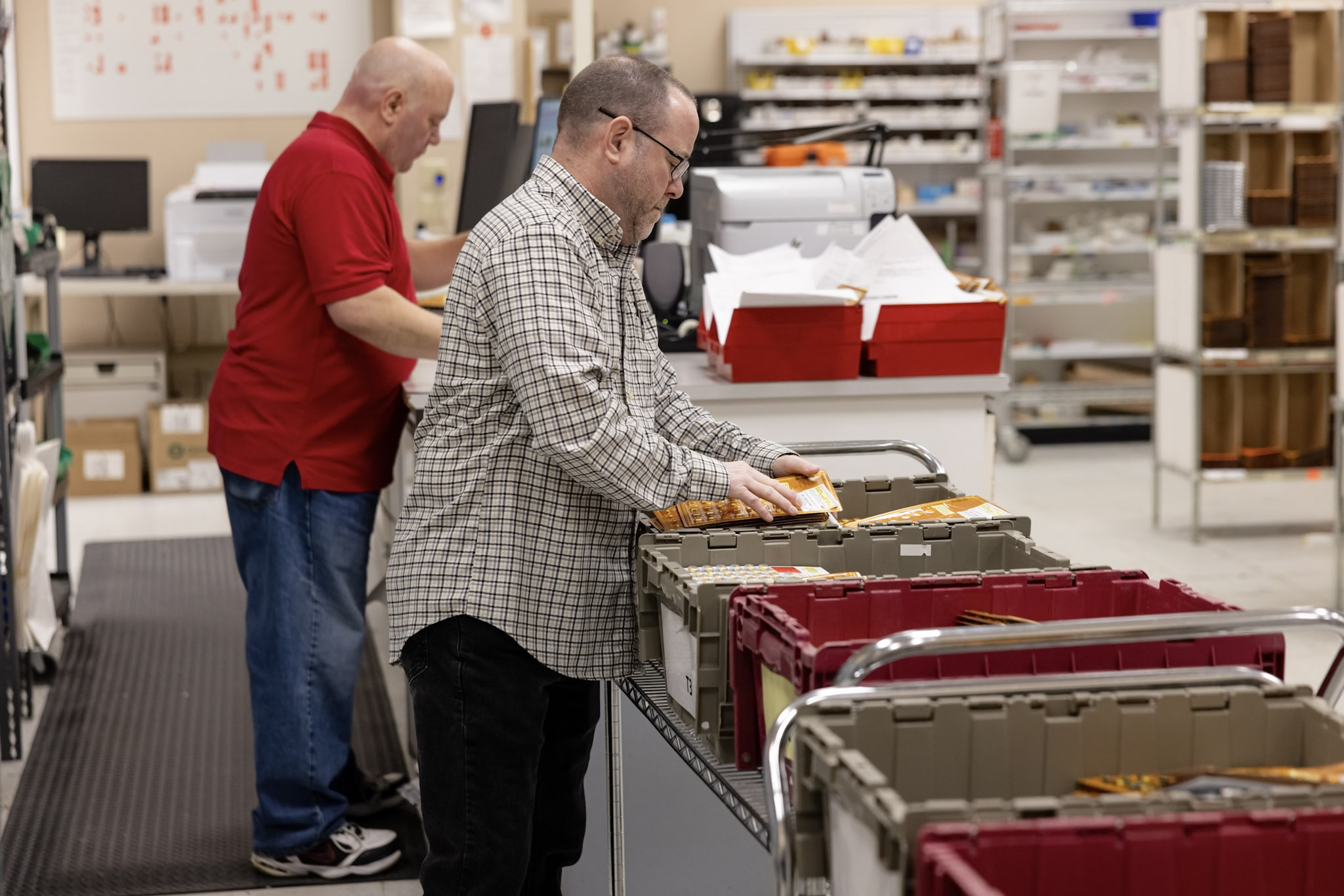 Two men work in a mailing and packaging area. One, wearing a red shirt, stands at a computer workstation. The other, in a checkered shirt, organizes materials into bins on a cart. Shelves filled with supplies are visible in the background.