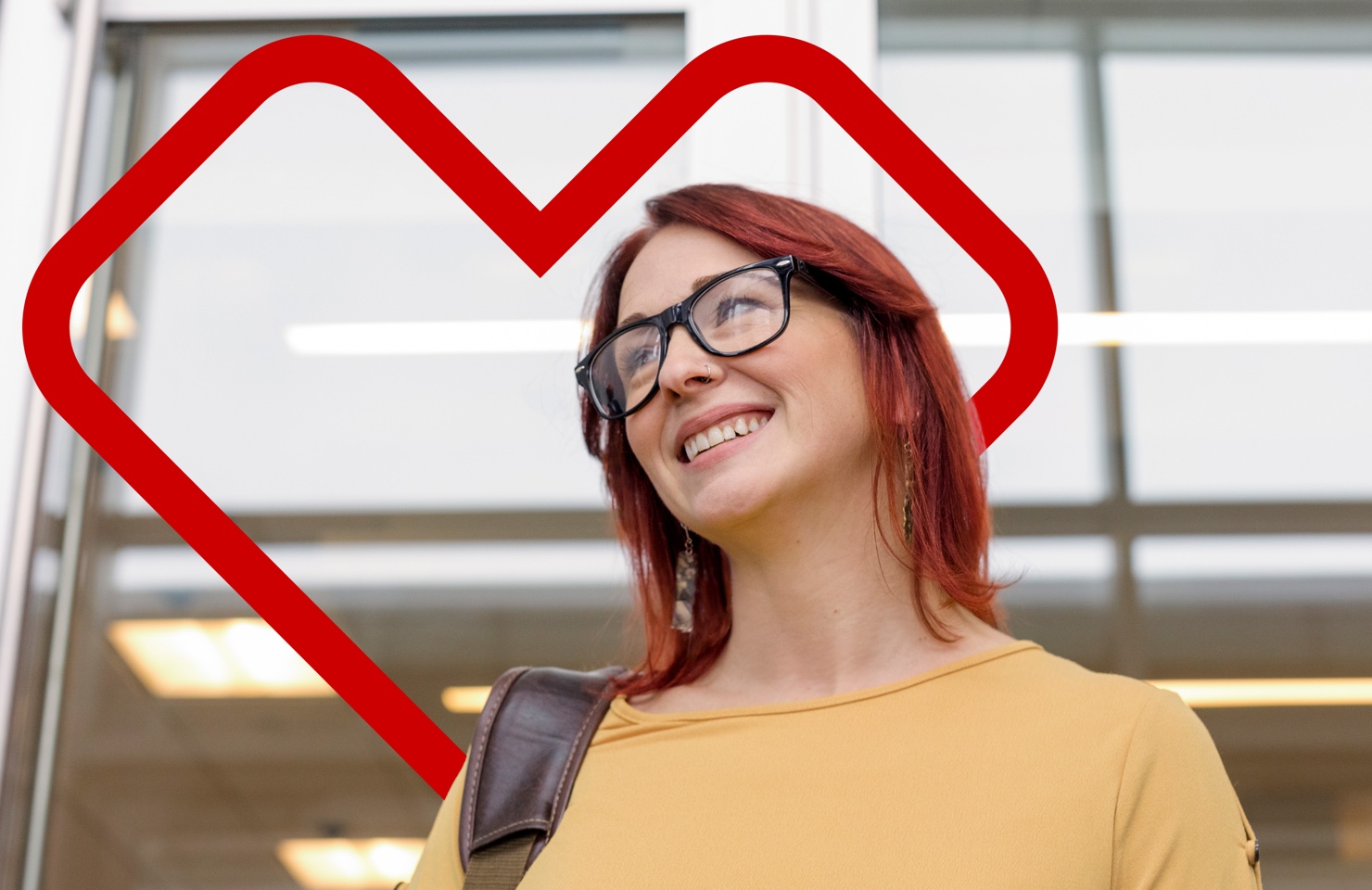 A person with red hair and glasses is smiling while standing in front of a large window. They are wearing a yellow top and a brown shoulder bag. A red heart outline frames their face in the background.