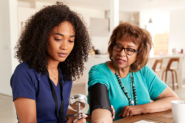 A young healthcare worker with curly hair in a blue uniform measures the blood pressure of an older woman wearing glasses and a turquoise shirt. They are seated at a table in a well-lit room with a modern kitchen in the background.