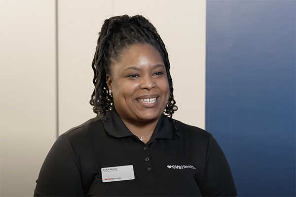 A person with braided hair, wearing a black CVS Health polo shirt and name tag, smiles while sitting in a well-lit room with a white wall on the left and a blue wall on the right.