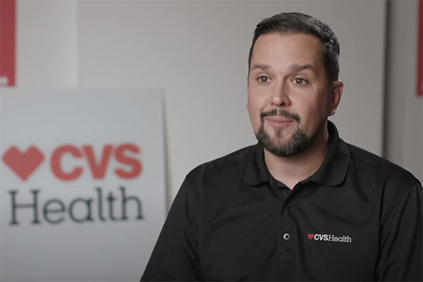 A man with short dark hair and a beard is wearing a black shirt with the CVS Health logo. He is sitting in an office setting, and there is a CVS Health sign visible in the background.