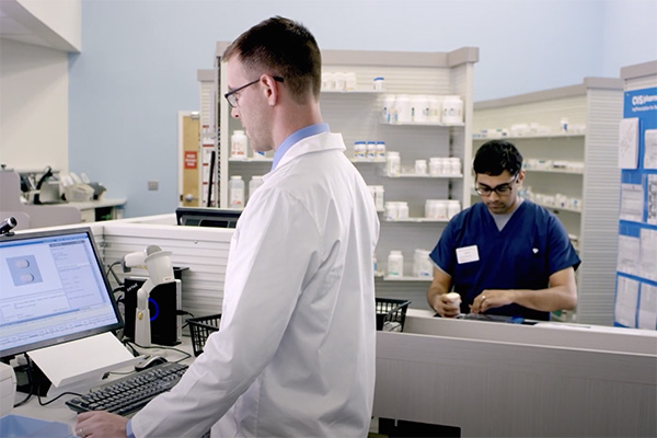 Two pharmacists are working in a modern pharmacy. One, wearing a white lab coat, is using a computer at a desk in the foreground. The other, in navy scrubs, is organizing medication bottles on a shelf in the background. Shelves with pharmaceuticals are visible.