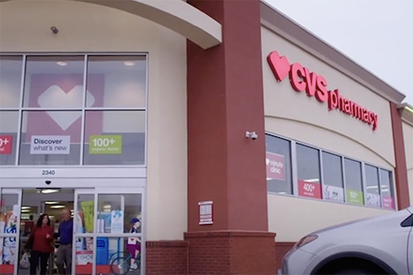 A CVS Pharmacy storefront with large glass windows and a red and white CVS Pharmacy sign above the entrance. Various promotional posters are displayed on the windows, and a car is parked in front of the entrance. People are visible exiting the store.