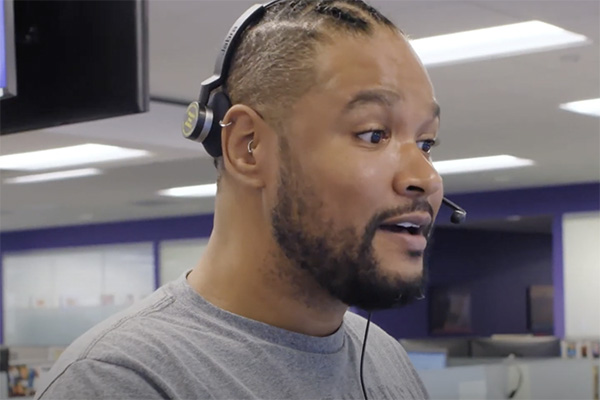 A man with braided hair and a beard is wearing a headset with a microphone. He is looking slightly to the right and appears to be in an office setting with bright overhead lighting and blurred background elements.