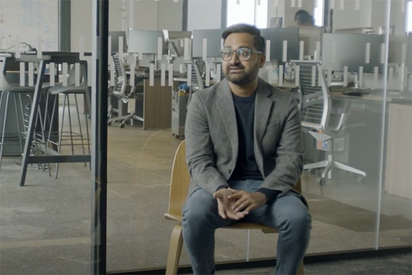 A man with glasses, dressed in a blazer and jeans, is seated on a wooden chair in a modern office setting. He is looking slightly to the left, with several workstations and office chairs visible behind a glass partition in the background.