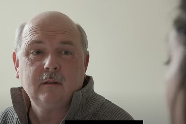 A mature man with a balding head and mustache is looking off to the side. He is wearing a beige sweater with the collar slightly unzipped. The background is plain and light-colored, and part of another person's face is visible in the foreground.