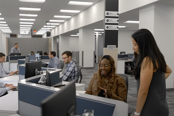 A modern office with several employees working at desks. A woman in a brown shirt is seated, engaging in conversation with a standing woman in a gray dress. The office is well-lit with a minimalistic design and an open workspace layout.