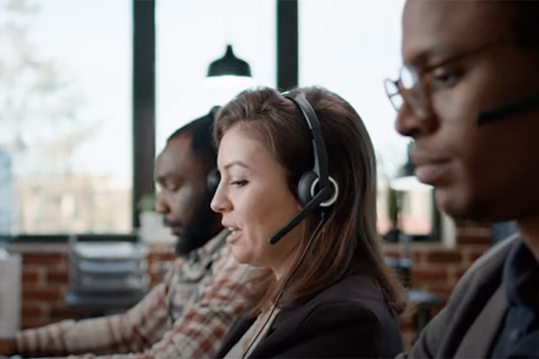 Three people are working at desks with headsets on in a modern office environment. They are focused on their tasks, possibly engaged in a call center or customer support setting. The office has large windows in the background letting in natural light.