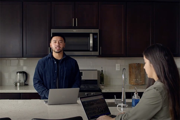 A man stands in a kitchen with dark wooden cabinets, speaking while using a laptop on the counter. A woman sits at the kitchen island with her back turned to the man, also using a laptop. The kitchen features a microwave, stove, and kettle.