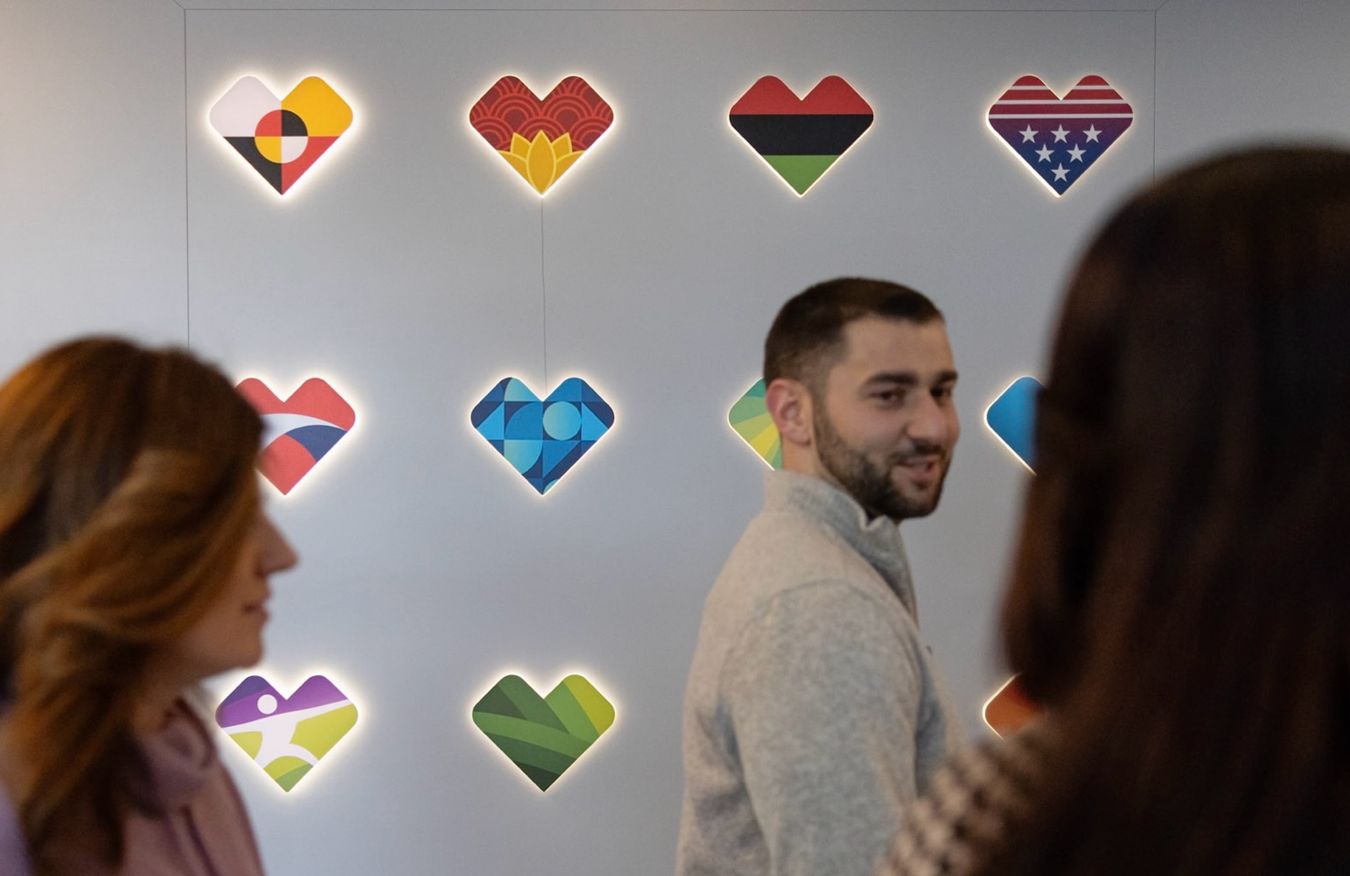 Three people in conversation in front of a wall featuring brightly colored heart-shaped symbols, each with a unique design representing different identities and themes. The hearts are lit, drawing attention to their vibrant colors.