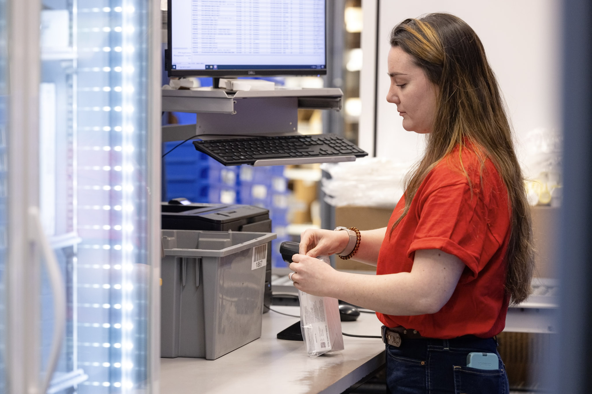 A woman wearing a red shirt scans a package at a workstation with a computer and gray storage bins. She appears to be processing the package, with shelves and additional bins visible in the background.