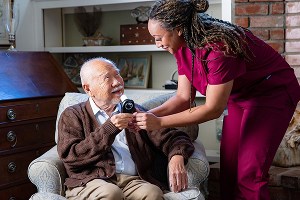 A senior man sits in an armchair, smiling as he holds a small black device handed to him by a nurse wearing a maroon uniform. The background includes a wooden cabinet and shelves with decorative items. Both the man and the nurse appear happy and engaged.