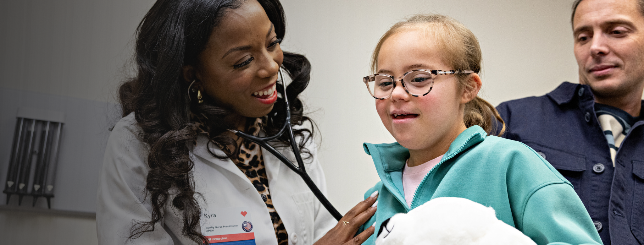Clinician using a stethoscope to examine a child in a clinic exam room while an adult stands nearby, with medical equipment visible on the wall.