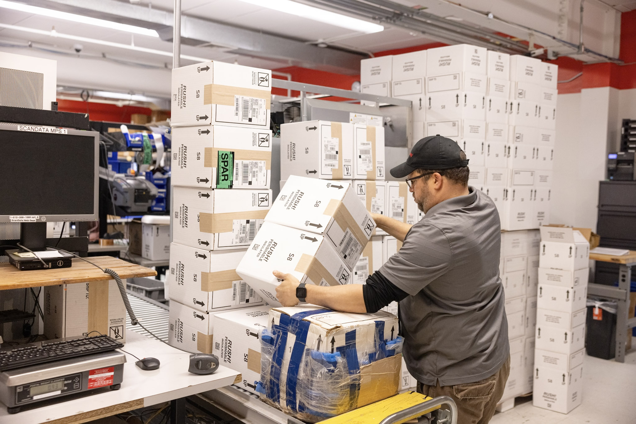 A worker wearing a gray shirt and black cap places a box onto a stack of packages in a warehouse filled with shelves of similar boxes. In the foreground, there's a workstation with a computer, scanner, and other packing equipment.