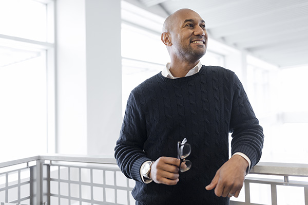 A person with a clean-shaven head, wearing a navy blue sweater and white collared shirt, stands by a railing in a bright, modern space. The person is smiling and holding eyeglasses in their hand, looking off to the side.