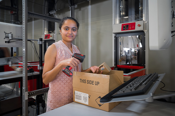 A woman stands at a work station, smiling and using a handheld barcode scanner on a cardboard box labeled "This Side Up." She is surrounded by industrial equipment and work tables, with a computer keyboard mounted on an adjustable arm to her right.