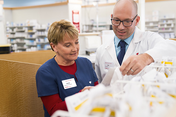 Two pharmacists, a woman in a blue uniform and a man in a white coat, are examining medication labels together in a pharmacy. Shelves filled with medication are visible in the background. Both are focused on their task.