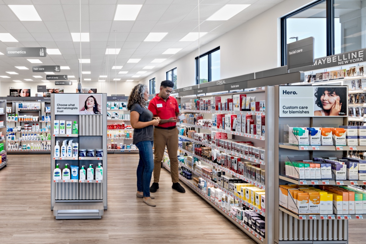 Two people stand in a brightly lit drugstore aisle, both looking at skincare products on a shelf. The person on the left is wearing a green shirt and jeans, and the person on the right is dressed in a red shirt and khaki pants, appearing to assist. Various skincare and beauty products are neatly arranged on shelves.