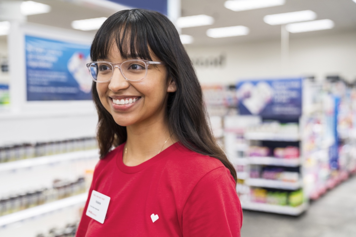 A smiling person with glasses and long hair wearing a red shirt and name badge stands in a brightly lit pharmacy aisle filled with various products and medicines.