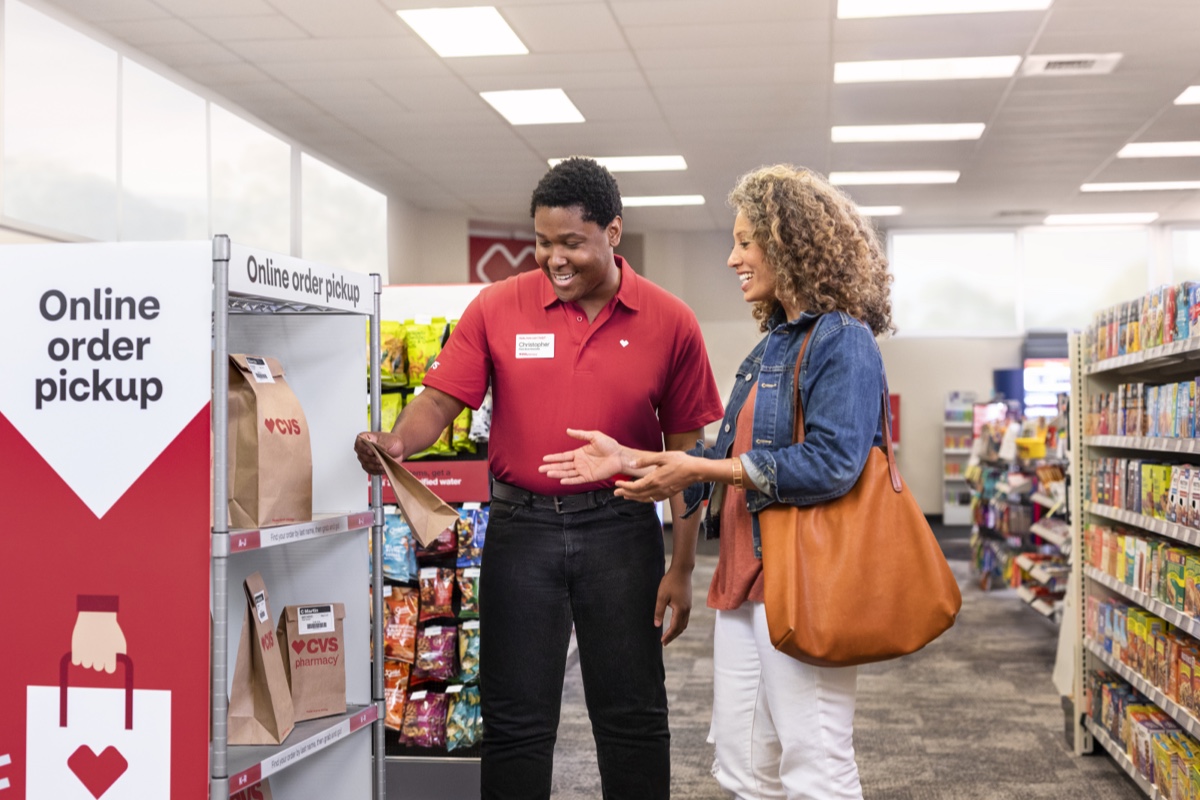 An employee in a red shirt and name tag assists a woman with curly hair and a brown tote bag in retrieving her online order from a pickup shelf in a store. The woman smiles as the employee hands her a bag. Shelves with various products are visible in the background.