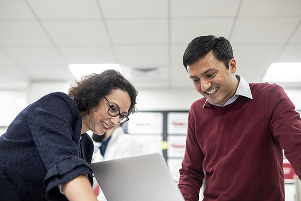 Two people are smiling and looking at a laptop screen in an office setting. One is wearing glasses and a dark blazer, and the other is in a red sweater with a white shirt underneath. They appear to be collaborating or discussing something on the computer.