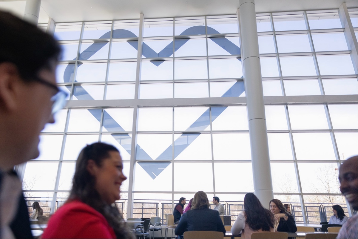 A group of people sits and converses in a bright indoor space with large windows. Visible in the background is a structural design on the windows forming the shape of a heart. The atmosphere appears casual and social, with natural light flooding the area.