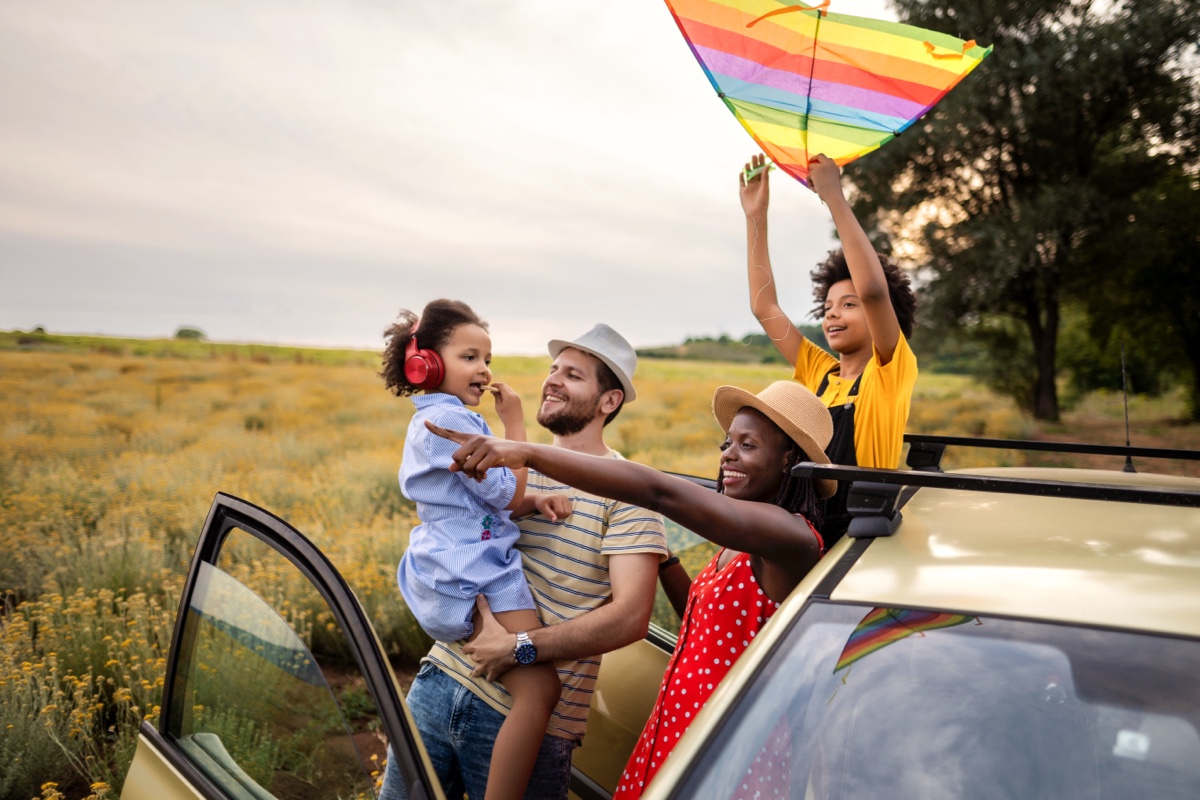 A happy family of four enjoys an outdoor day. The father, carrying a young girl wearing headphones, smiles at the mother, who is pointing. The older child, standing on the car's roof, holds a colorful kite. They are surrounded by a blooming meadow.