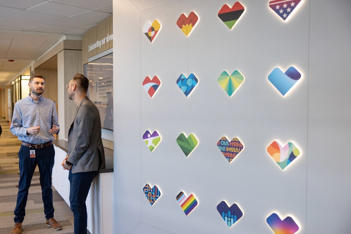 Two men are standing and conversing in a hallway next to a white wall featuring a display of various colorful heart-shaped designs with different flags, symbols, and patterns. Different offices and rooms are visible down the hallway.