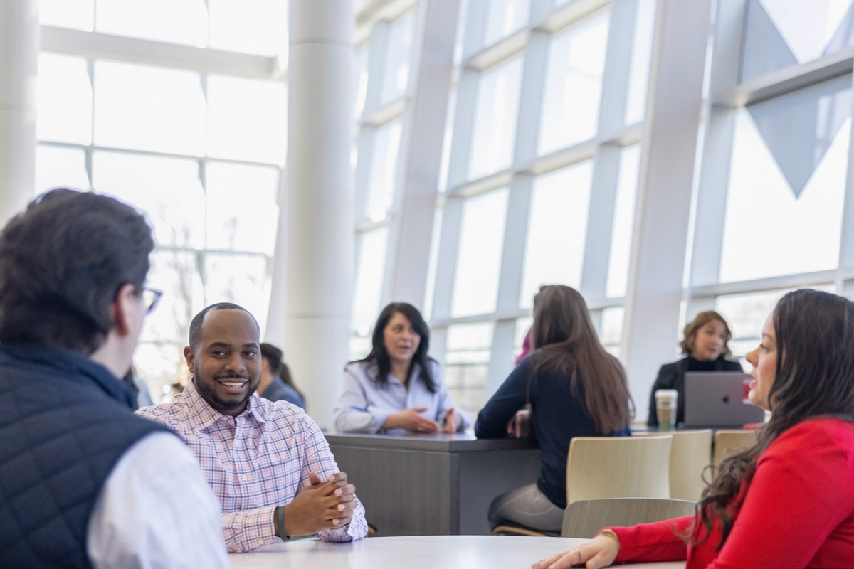 A group of people sits at tables in a modern, bright office space with large windows. In the foreground, three individuals engage in conversation around a table. Other people work and chat at tables in the background.