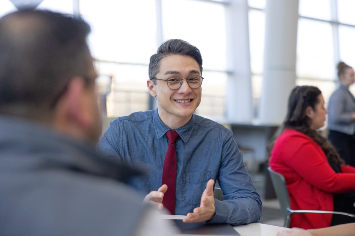 A person wearing glasses, a blue dress shirt, and a red tie is smiling and gesturing with their hands while sitting at a table. Another person with blurred features is seated opposite them. The background features large windows and other people engaged in conversation.