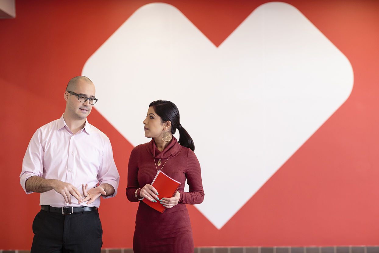 Two people are standing in front of a large white heart on a red background. The person on the left, wearing glasses and a light pink shirt, is speaking, while the person on the right, in a maroon dress holding a red notebook, listens attentively.