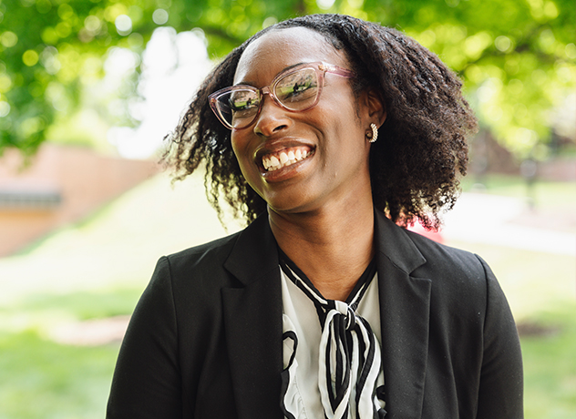 A person wearing glasses, a black blazer, and a white blouse with a black and white scarf stands outdoors. They are smiling and have shoulder-length curly hair. The background is a green, sunlit area with trees and grass.