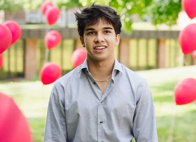 A young man with dark hair and a light grey button-up shirt stands outdoors, surrounded by red balloons. The background includes a green lawn, trees, and a building. He is smiling slightly and looking ahead.