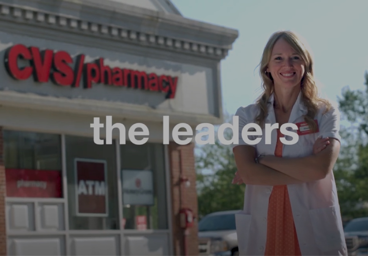 A smiling pharmacist stands in front of a CVS Pharmacy. She is wearing a white lab coat and has her arms crossed. The building has the CVS Pharmacy logo on it. The text "the leaders" is written across the image. An ATM is visible in the background.